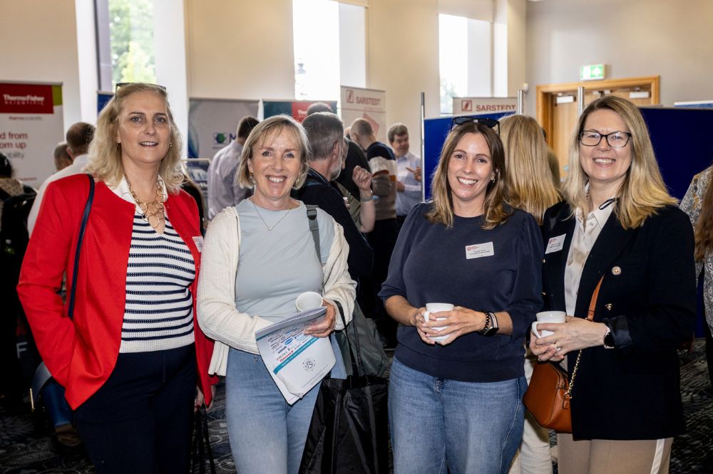 Four female delegates posing for a picture in front of stands and other delegates milling around in an exhibition space in a large white windowed room.
